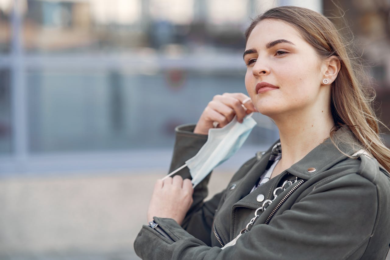 A woman removes her face mask outdoors, exuding confidence and style in a casual jacket.